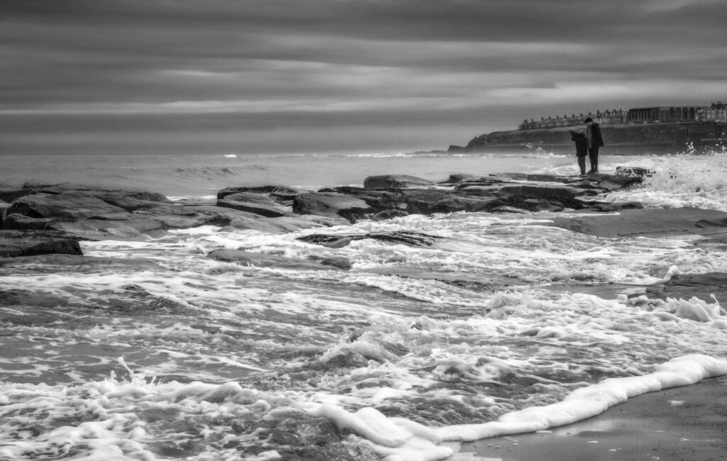 Monochrome scene of a rocky coastline with waves and two people.